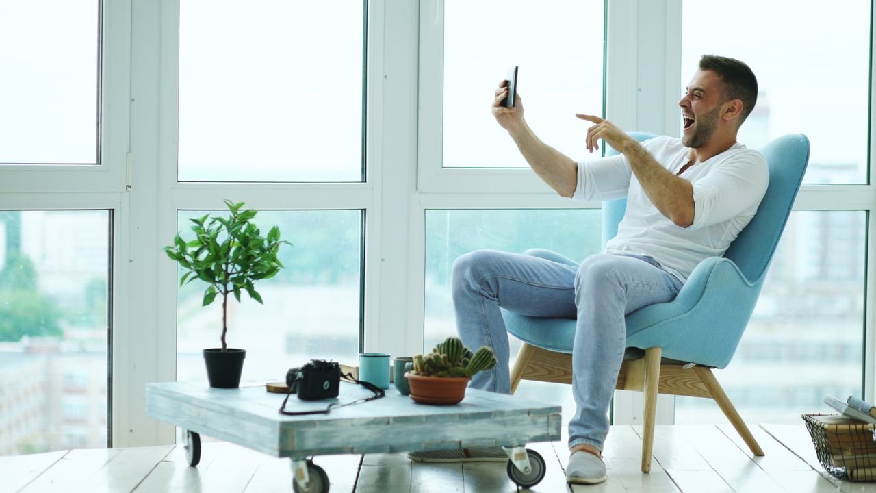 The Art of Drawing Readers In: Your attractive post title goes here A cheerful man sitting in a modern living room, enjoying a video call on his smartphone with a scenic city view.